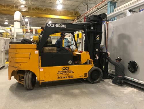 CCI Industrial Constructors - A forklift operator carefully maneuvers a large yellow forklift while a coworker assists with guidance for handling heavy machinery in an industrial setting.