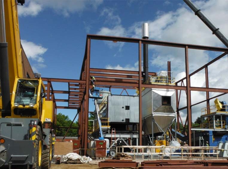 CCI Industrial Constructors - A construction site with structural steel framing in progress, large crane on the left, and various building materials scattered on the ground under a cloudy sky.