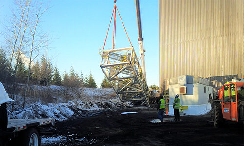 CCI Industrial Constructors - Industrial workers in high-visibility jackets using a crane to lift a large metal structure onto a truck at a construction site, with a snowy background indicating cold weather conditions.
