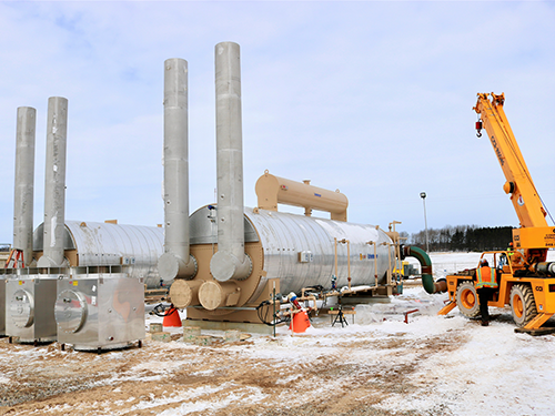 CCI Industrial Constructors - Construction site in a cold environment with industrial piping and machinery, including a large tank and a mobile crane operating nearby, set against a backdrop of a partially snow-covered ground and a clear sky.