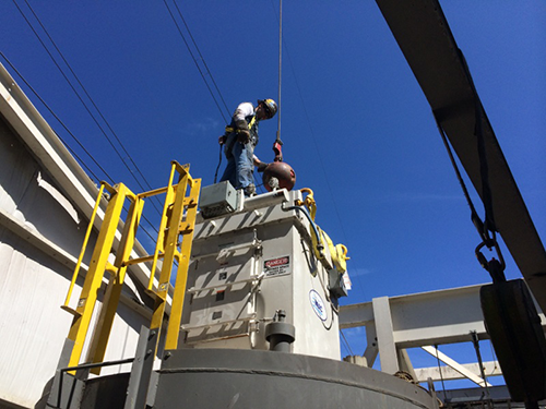 CCI Industrial Constructors - Worker in a harness performing maintenance or inspection on industrial equipment outdoors under a clear blue sky.