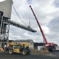 CCI Industrial Constructors - A large red mobile crane hoisting equipment next to a mining conveyor structure, with construction and dump trucks nearby, under a cloudy sky.