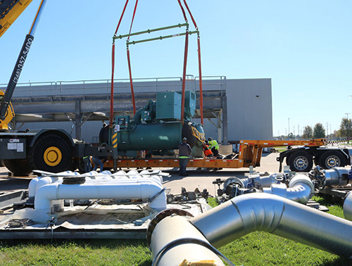 CCI Industrial Constructors - Industrial equipment installation: workers in high-visibility vests use a crane to carefully position a large green machine onto a transportation platform, with various pipes and components laid out on the ground, against an industrial facility backdrop under a clear blue sky.