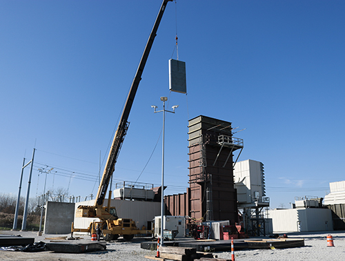 CCI Industrial Constructors - A mobile crane hoisting a large panel towards a multi-story structure at an industrial construction site under a clear blue sky.