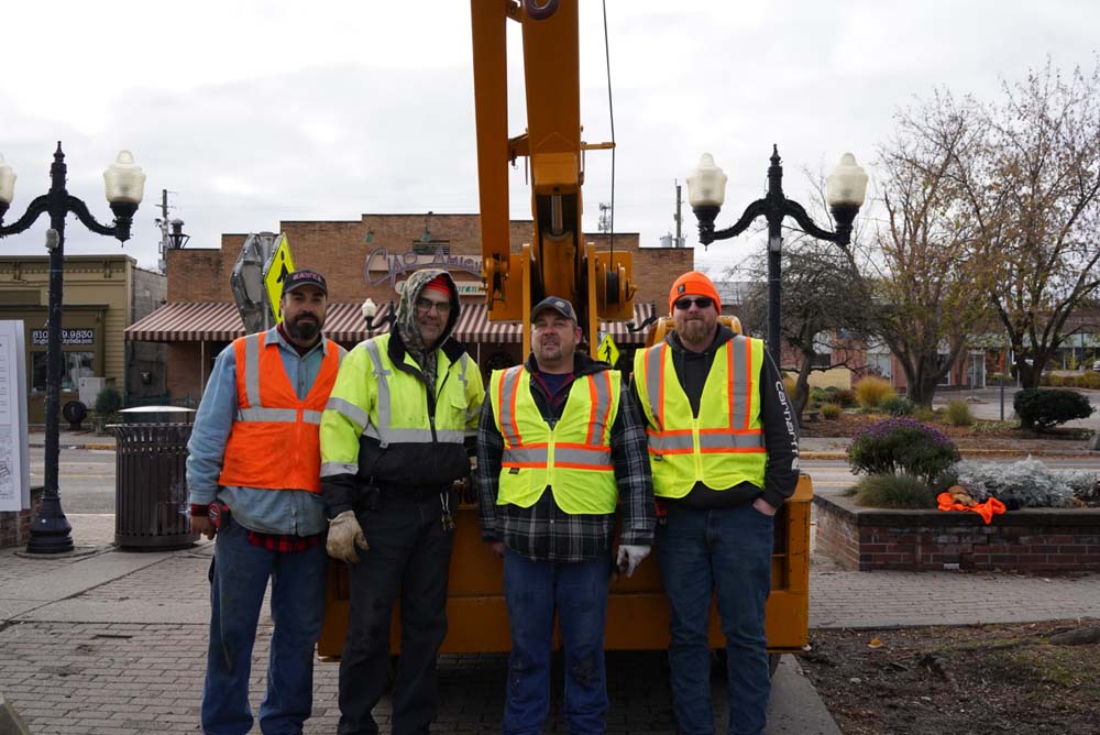 CCI Industrial Constructors - A group of four construction workers posing together in front of a piece of heavy machinery, dressed in safety gear with high visibility vests, on a cloudy day in an urban setting.