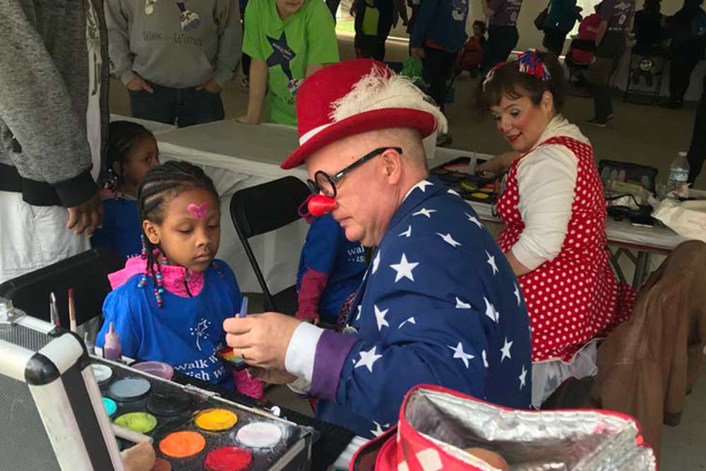 CCI Industrial Constructors - A clown in a star-spangled outfit paints the face of a young girl at a community event while another person in a polka-dot costume looks on.