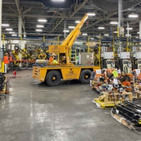 CCI Industrial Constructors - An industrial scene with a mobile crane in operation at a busy manufacturing plant, where workers with safety vests are engaged in various tasks amidst machinery and equipment.