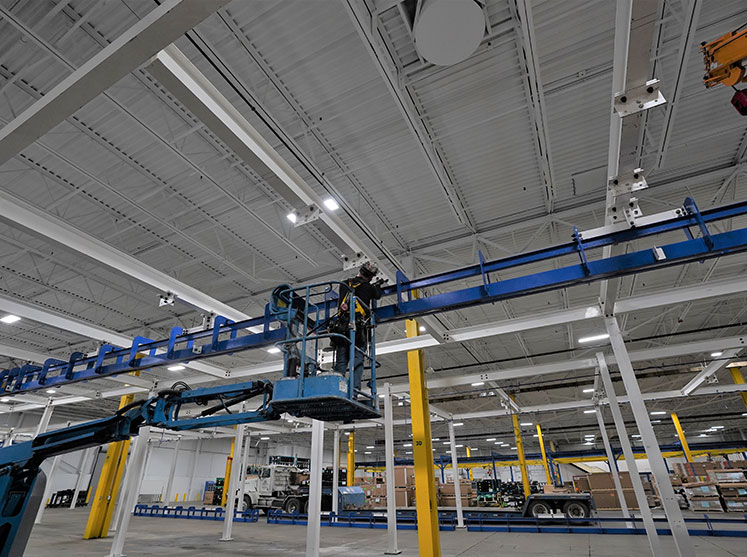 CCI Industrial Constructors - A worker on a blue scissor lift performing maintenance or installation services on overhead structures inside an industrial facility with high ceilings and bright lighting.