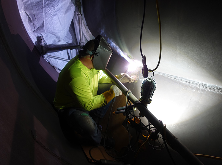 CCI Industrial Constructors - A skilled welder working with focus and precision in a tight, dimly lit space, the arc of the welding torch casting a bright glow on the metal surface.