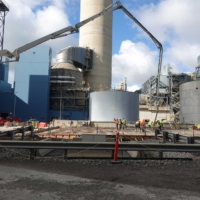 CCI Industrial Constructors - Construction workers collaborate on a large industrial site, with concrete pouring from a boom pump into formwork amidst towering structures and storage tanks under a cloudy sky.