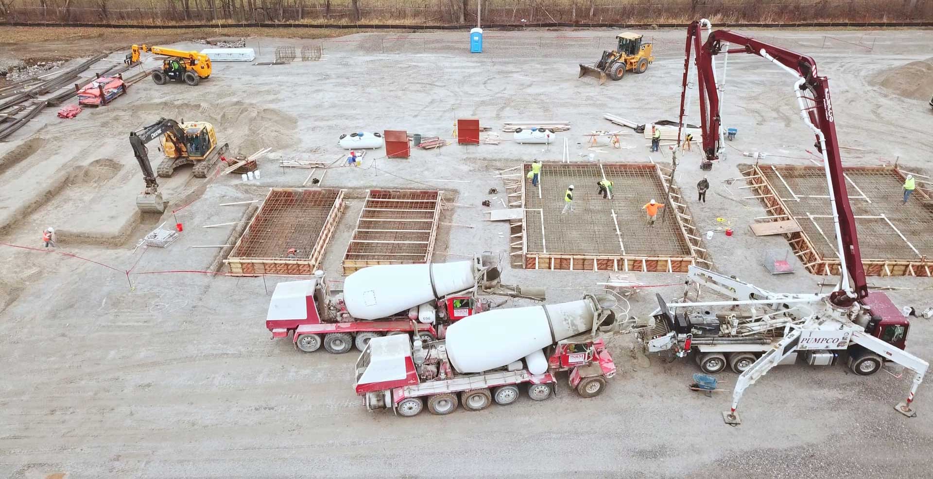 CCI Industrial Constructors - A busy construction site with workers collaborating on the foundation, as concrete is being poured from a pump connected to mixer trucks, amidst a backdrop of earthmoving equipment.