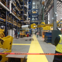 CCI Industrial Constructors - A worker in a hard hat and high-visibility vest attentively observes a large piece of cylindrical machinery being transported by an overhead crane inside an industrial manufacturing plant.