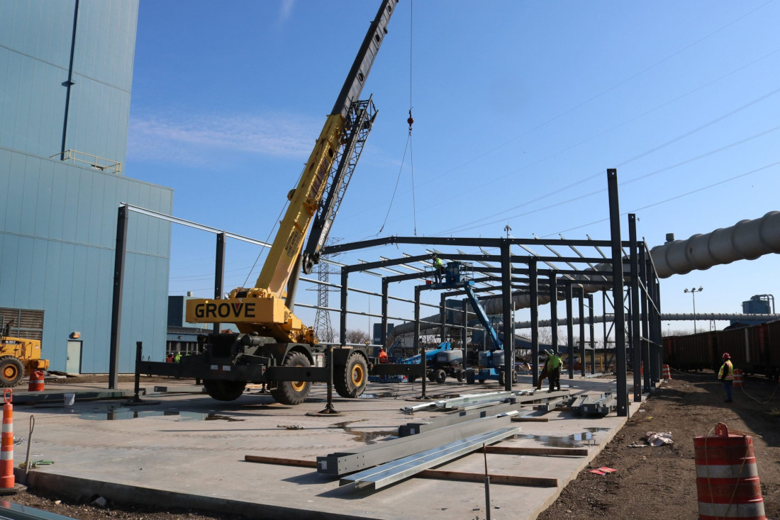 CCI Industrial Constructors - Industrial construction site with a mobile crane lifting materials for the assembly of a steel framework of a new building under a clear blue sky.